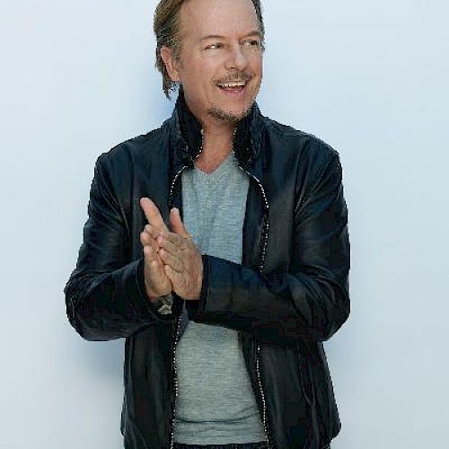 A man with a leather jacket and jeans is smiling and clapping hands against a white background.