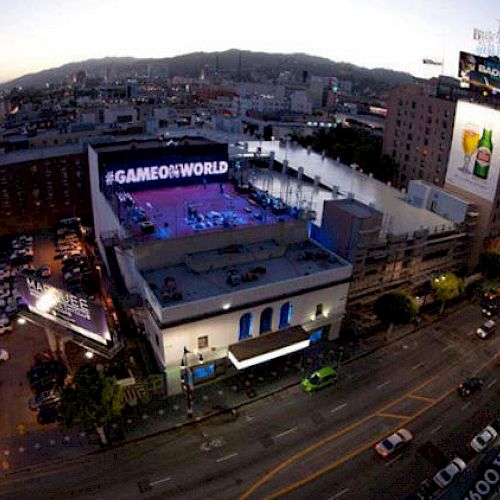 Aerial city view at dusk with buildings, streets, and a billboard reading #GAMEONWORLD.