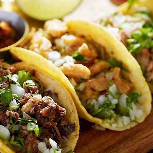 The image shows three tacos with meat, onions, cilantro, on a wooden board, with lime and sauce on the side.