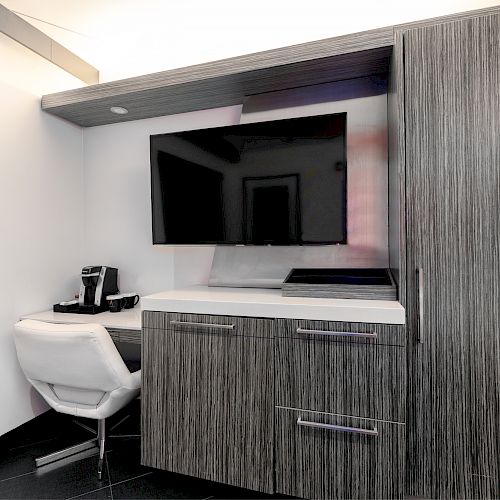 Modern kitchenette with dark wood cabinetry, white countertop, mounted TV, and a bar stool.