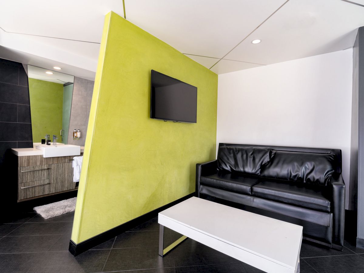A modern room with a black sofa, white coffee table, and a green accent wall featuring a TV. A small sink and mirror are visible.