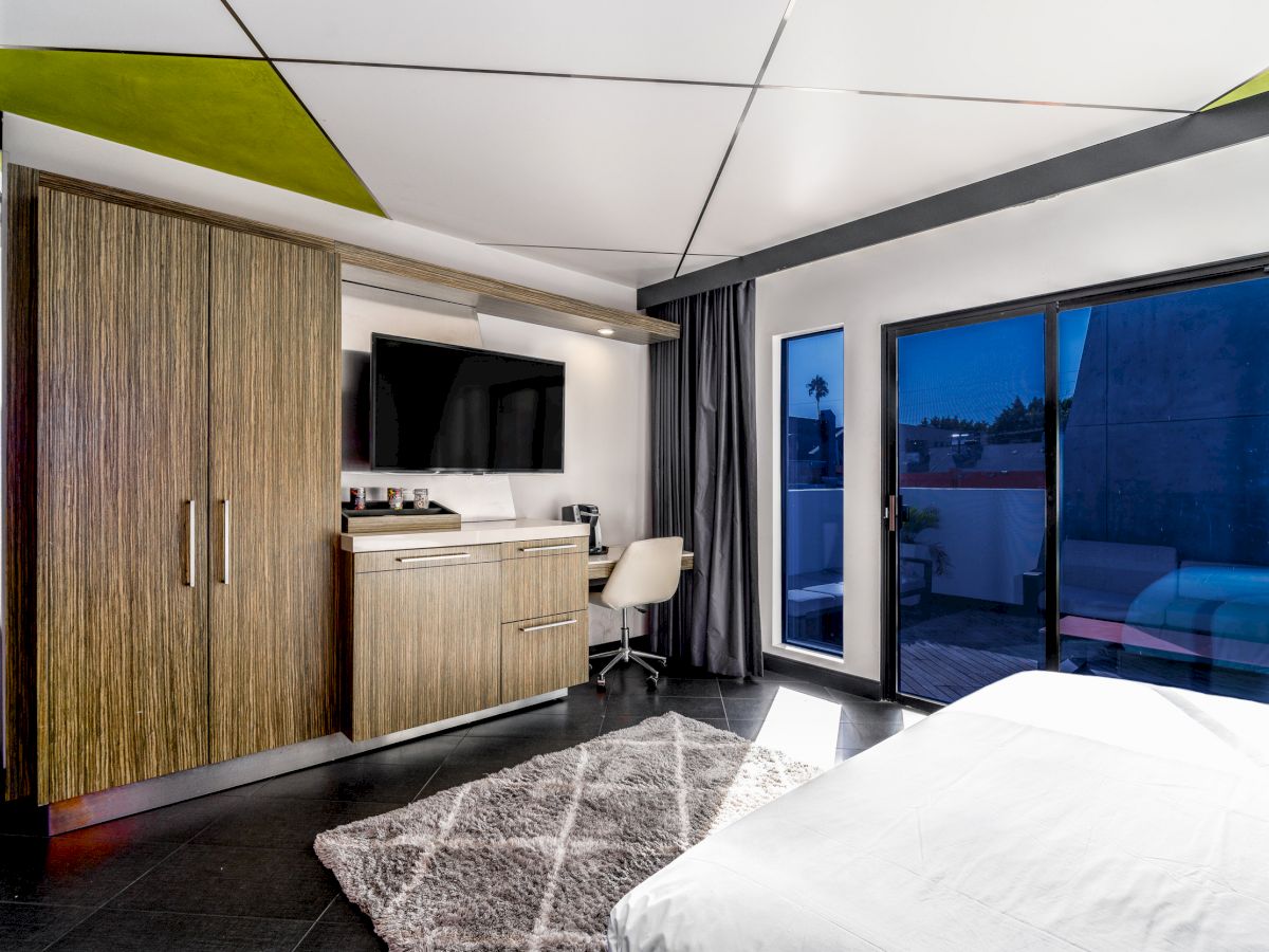 Modern hotel room with bed, desk, kitchenette, and balcony.